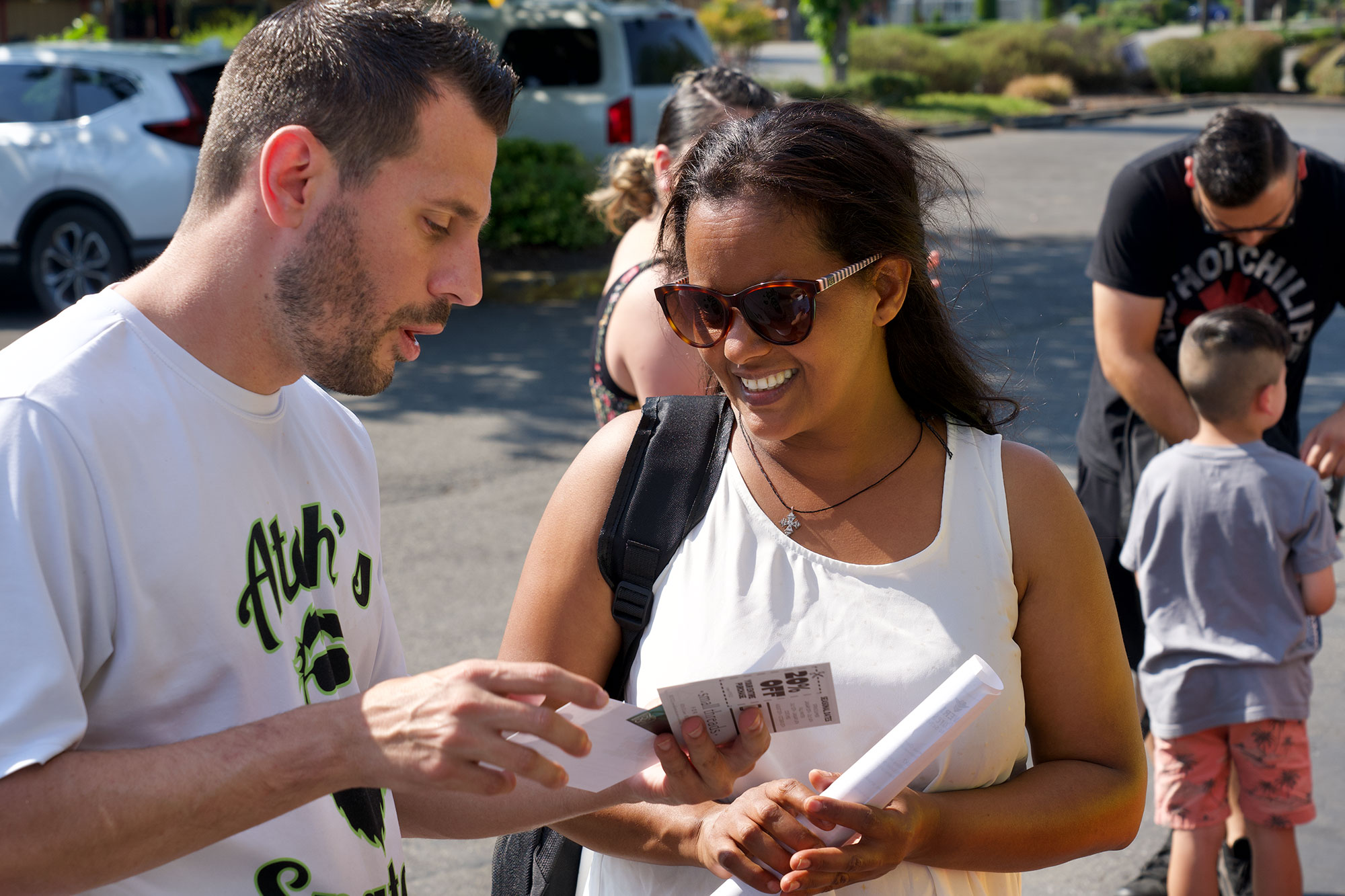 Man in white t-shirt talking to a woman with sunglasses and showing her a card